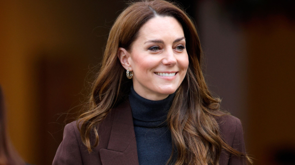 Kate Middleton attends an event in a turtleneck and brown jacket. She wears her hair long and in waves.